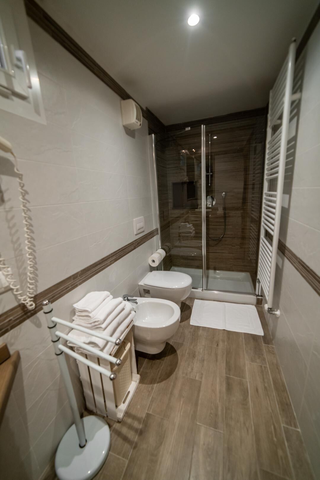 A modern bathroom with a glass-enclosed shower, a toilet, and a bidet. There is a towel rack with folded towels and a toilet paper roll.