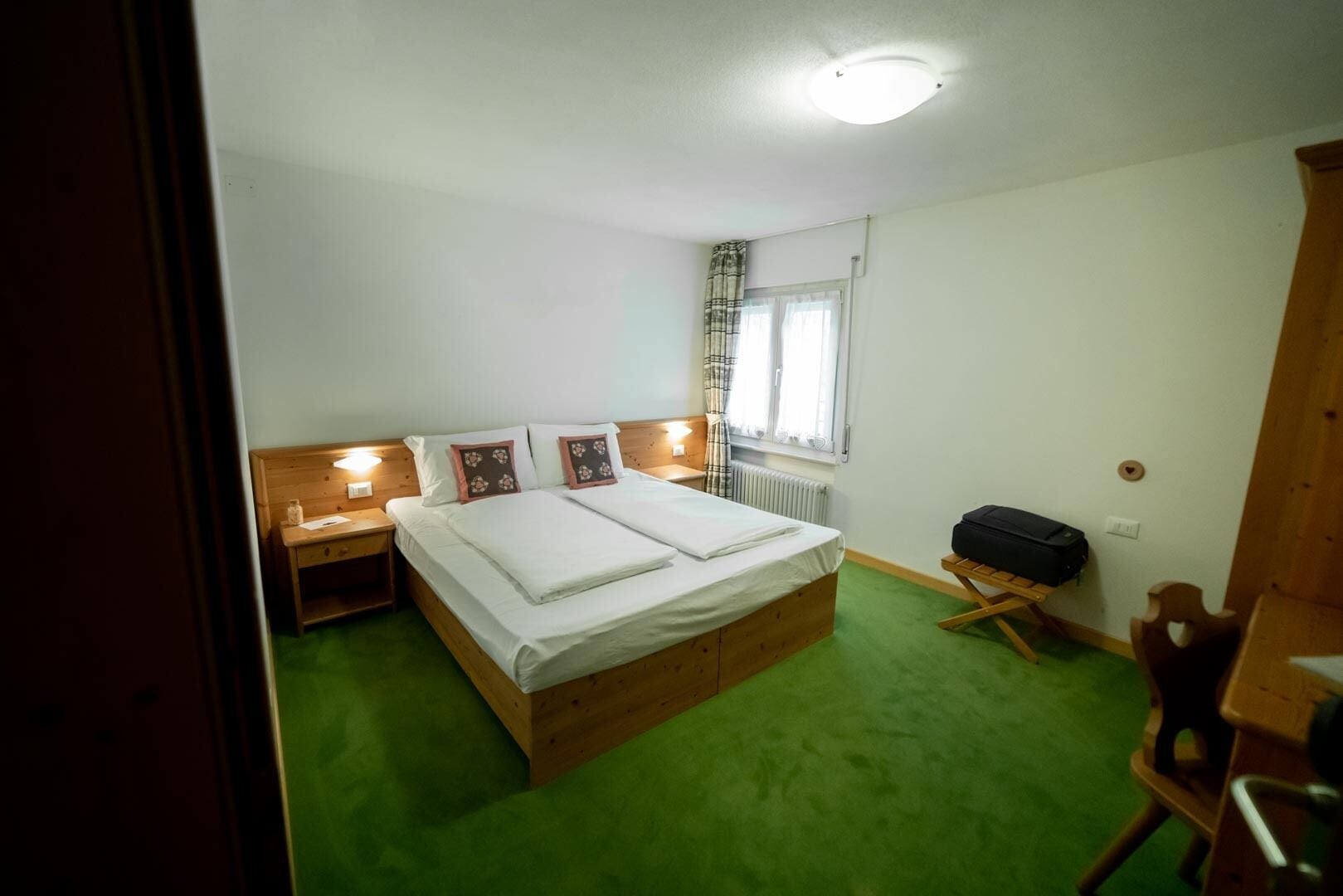 A room with a wooden bed, green carpet, a nightstand with a lamp, a suitcase, and a radiator.