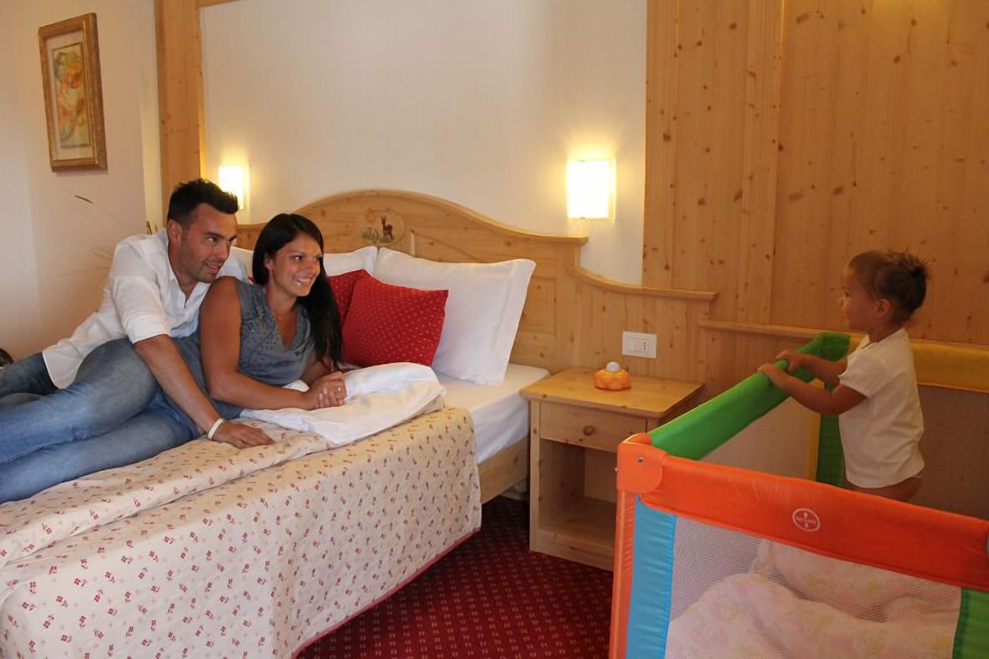 A hotel room with a wooden headboard, a bed with patterned bedding, a nightstand, and a small playpen.