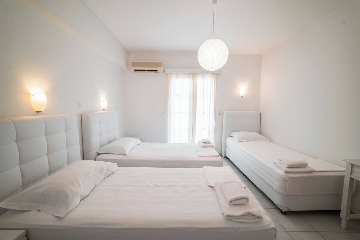 Quarto de hotel com duas camas, uma janela grande com cortinas, um lustre pendurado e um ar condicionado na parede.