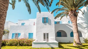 A white building with blue shutters, a palm tree, and a flower bed. - Hotel Sagterra (Naxos)