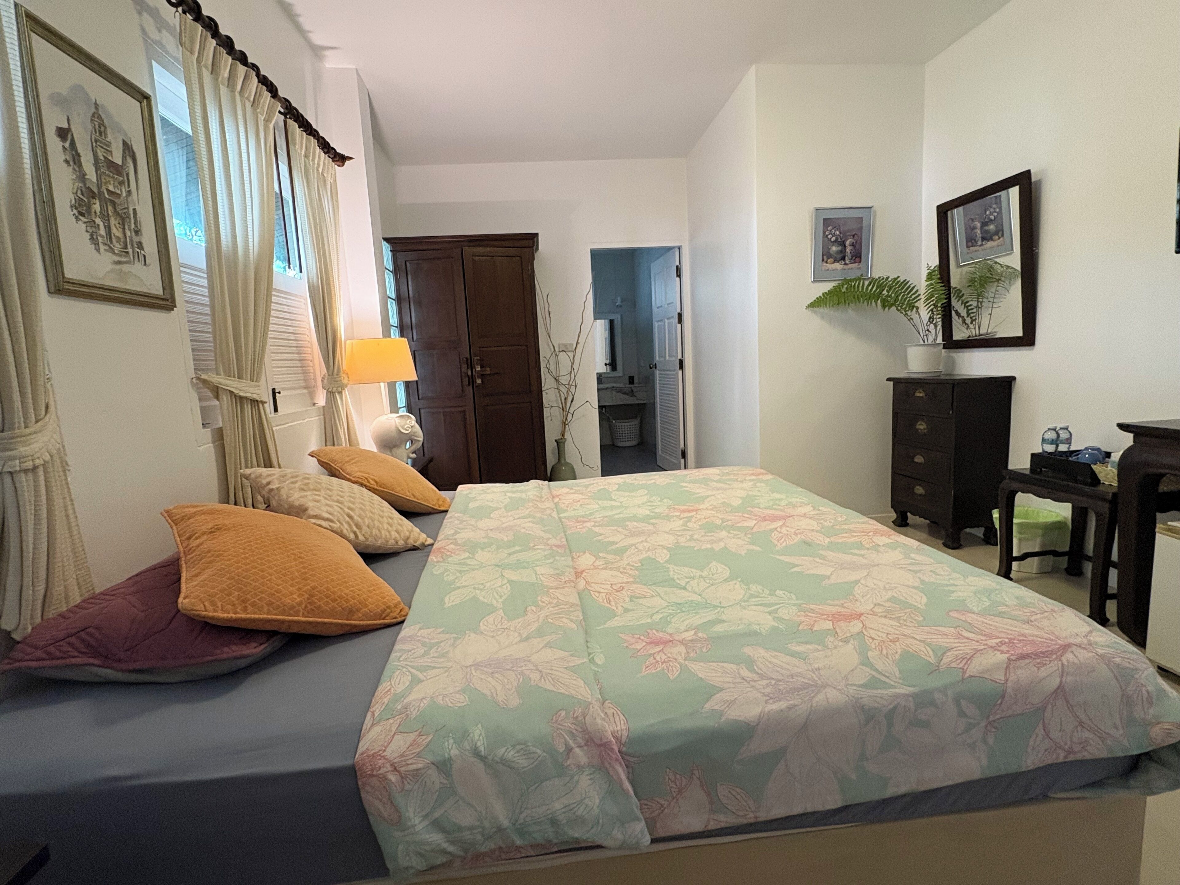 A bedroom with a bed, a dresser, a desk, and a chair. There is a window with curtains, a lamp, and a plant on the dresser.