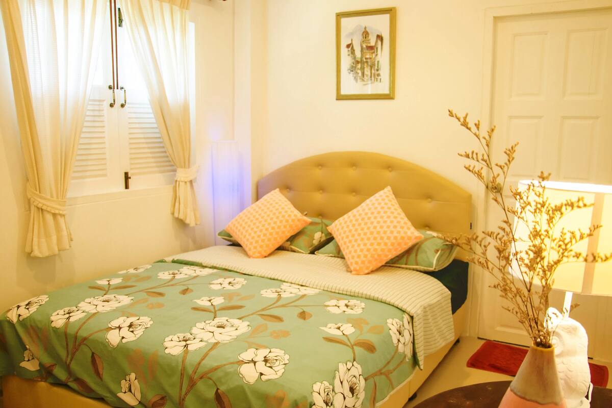 Un dormitorio con cama, mesita de noche, lámpara y ventana con cortinas.