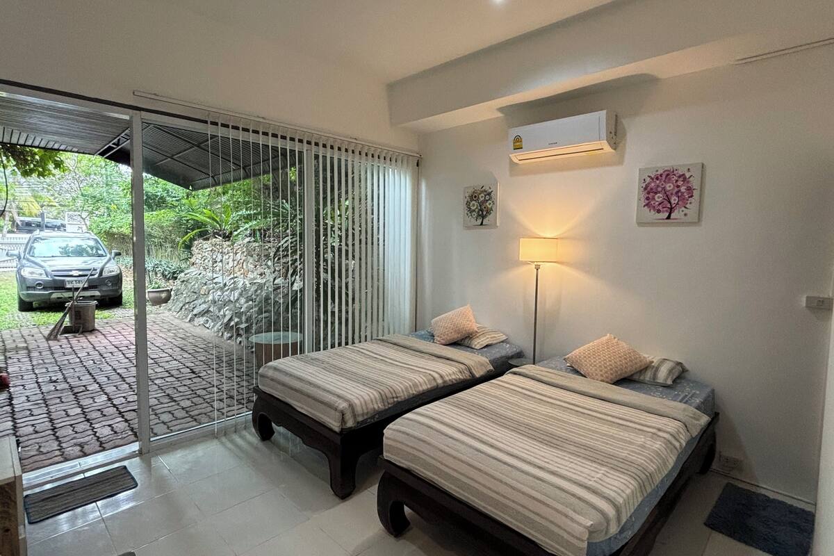 Habitación con dos camas, ventilador de techo, aire acondicionado instalado en la pared y vista a un coche estacionado afuera.