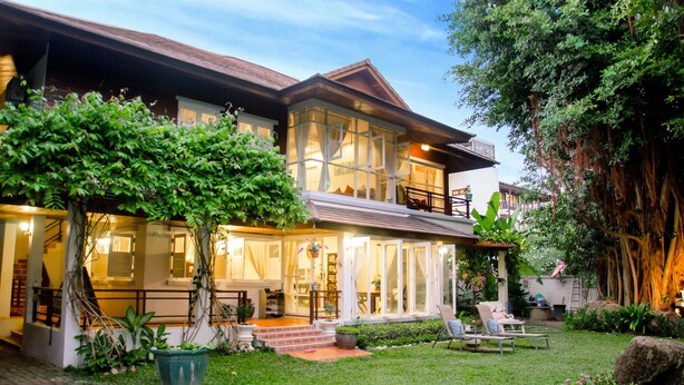 Une maison à deux étages avec une grande façade vitrée, un balcon et un jardin bien entretenu agrémenté d’un arbre et de plantes en pot.