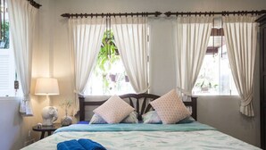 Une chambre à coucher avec un lit, deux fenêtres avec des rideaux, une table de chevet avec une lampe et une étagère fixée au mur, sur laquelle est posé un objet décoratif.