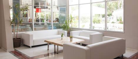 A modern interior with white sofas, a coffee table, and large windows offering a view of greenery.