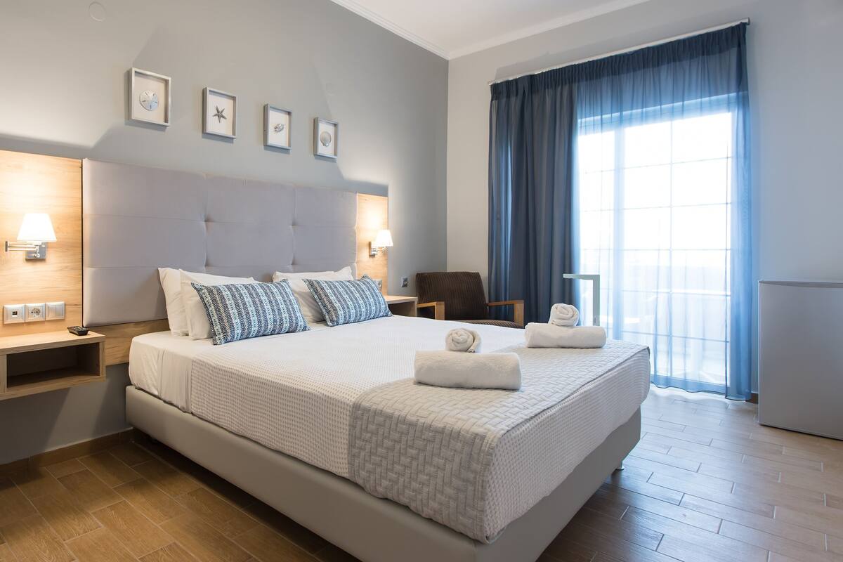 Quarto de hotel moderno com uma cama grande, mesas de cabeceira, uma cadeira, uma escrivaninha e uma janela ampla com cortinas.