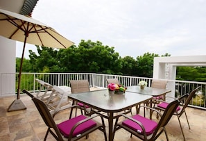 Um pátio com mesa de vidro, cadeiras de vime com almofadas rosa e um guarda-sol bege.