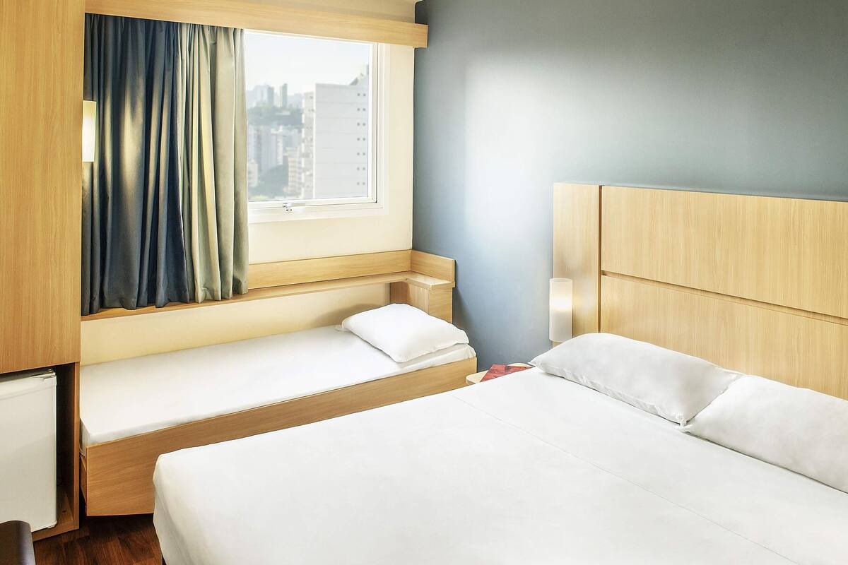 A hotel room with a bed, a window with curtains, a bedside table, and a lamp.