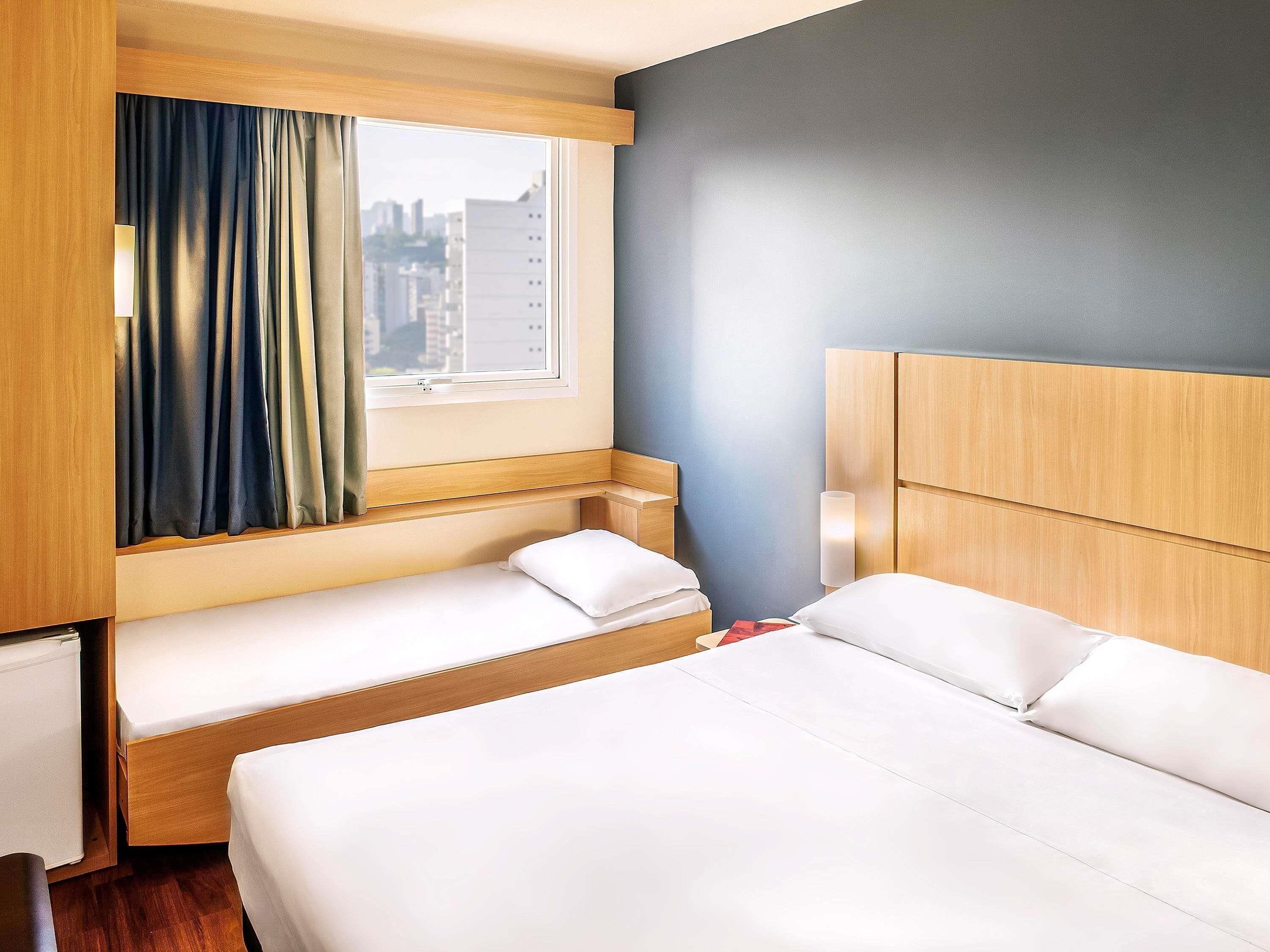A hotel room with a bed, a window with curtains, a bedside table, and a lamp.