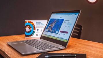 En bærbar computer på et træbord, der viser et hotelbookingsite, et visitkort og en notesbog.