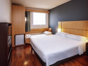 A hotel room with a bed, a desk, a small refrigerator, a window with curtains, and a nightstand with a lamp. - Ibis Belo Horizonte Savassi (Belo Horizonte)