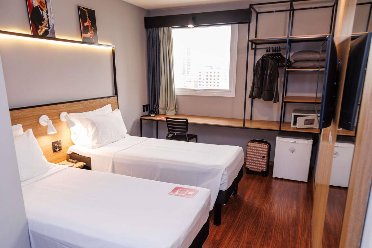 A hotel room with two beds, a desk, a chair, a wardrobe, a television, and a window with a city view.