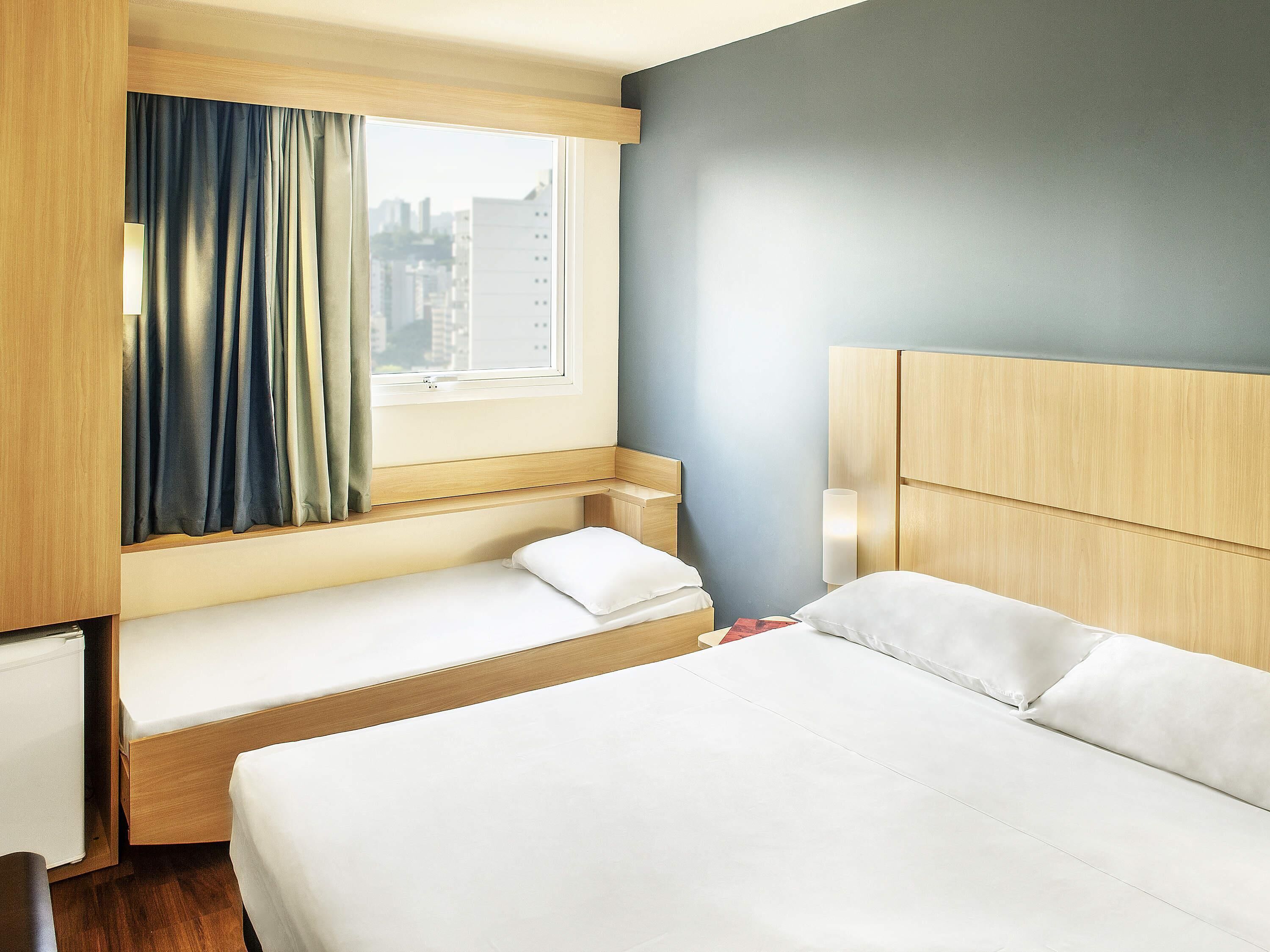 A hotel room with a bed, a window with curtains, a bedside table, and a lamp.