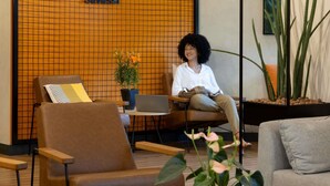 A woman sitting on a brown chair in a modern lobby with a grid-patterned wall, a laptop on a small table, and potted plants. - Ibis Belo Horizonte Savassi (Belo Horizonte)