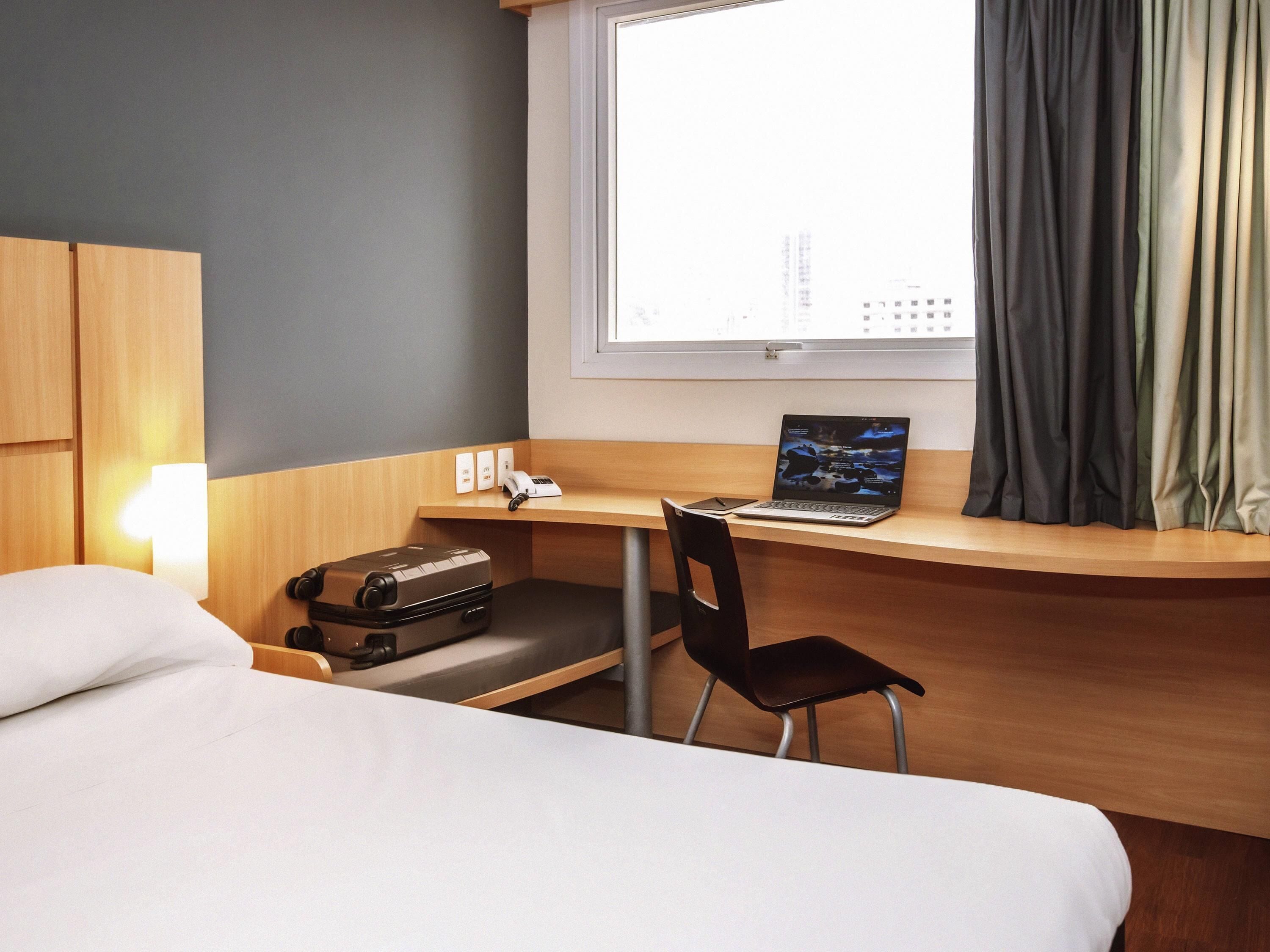 A hotel room with a bed, a desk with a laptop, a chair, a suitcase, and a window with curtains.