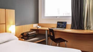 A hotel room with a bed, a desk with a laptop, a chair, a suitcase, and a window with curtains.