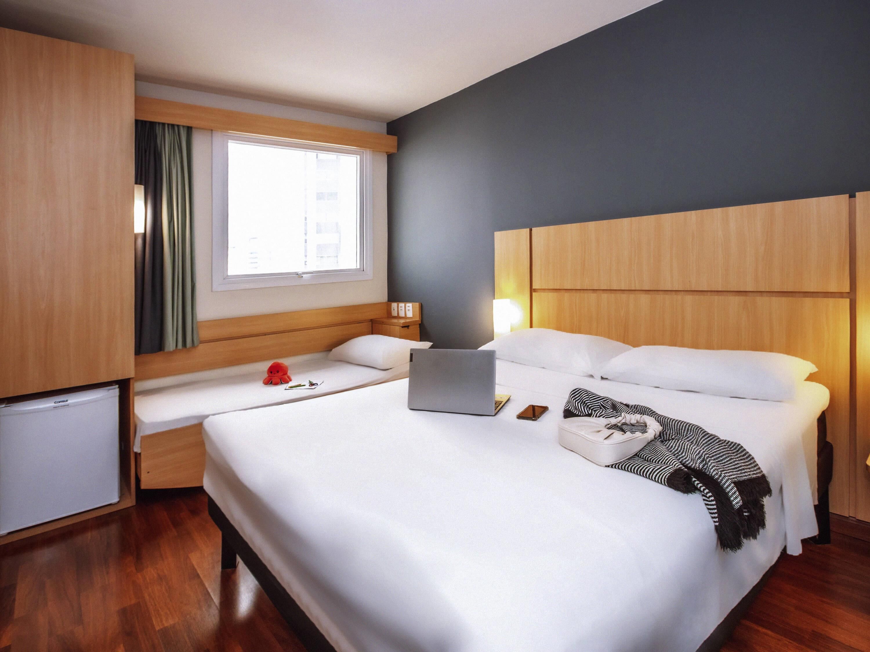 A hotel room with a bed, a desk, a laptop, a red toy, and a window with curtains.
