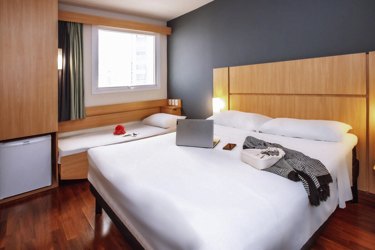 A hotel room with a bed, a desk, a laptop, a red plush toy, and a small refrigerator.