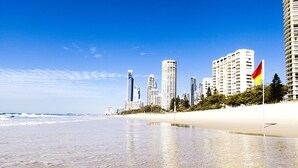 A beach with a sandy shore and ocean waves. There are tall buildings and a flag in the background. - Seacrest Beachfront Holiday Apartments (Surfers Paradise)