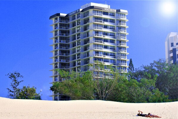 A high-rise hotel with multiple balconies, surrounded by trees and a sandy beach.