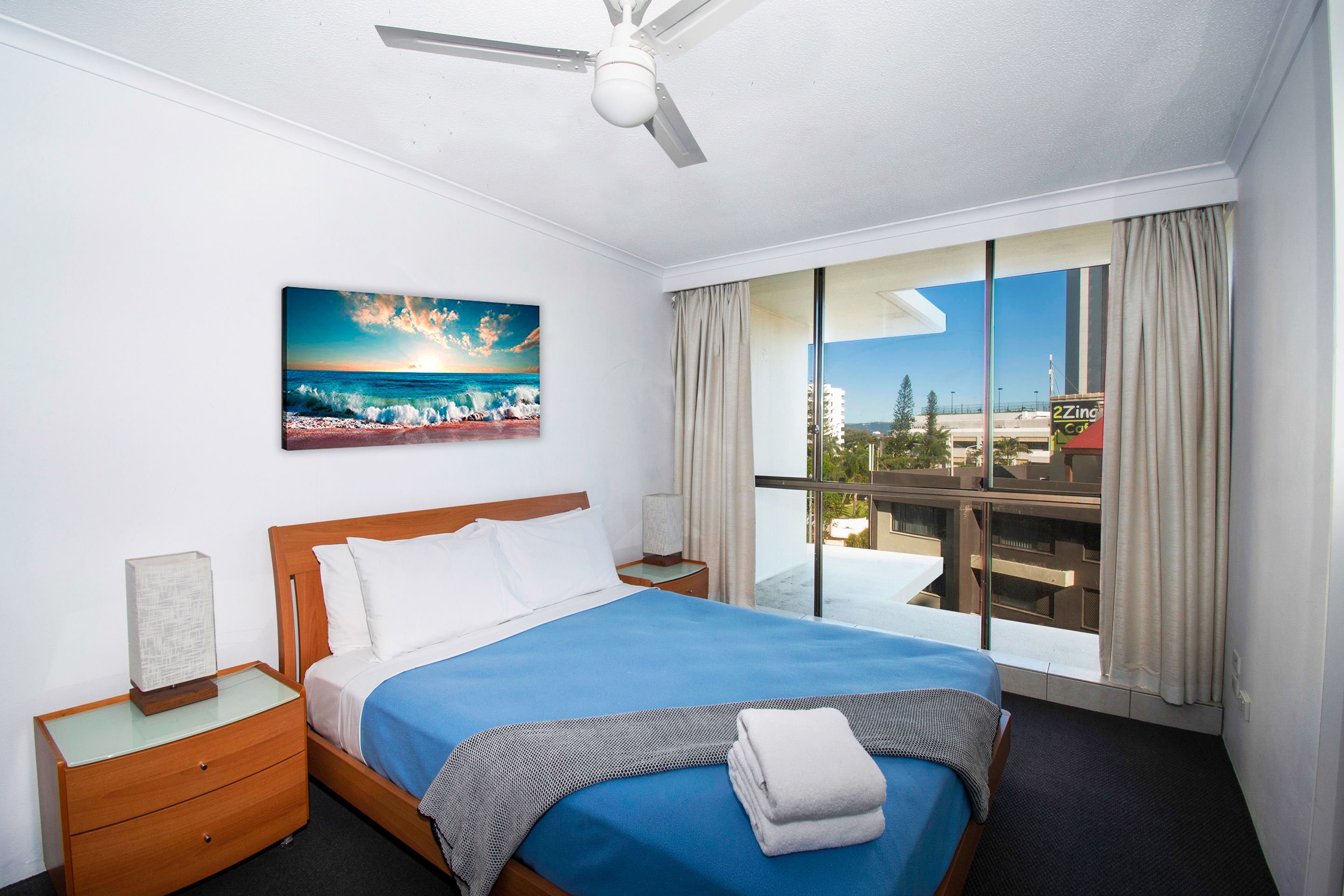 Quarto de hotel com cama, mesa de cabeceira, pintura de uma praia e varanda com vista para a cidade.