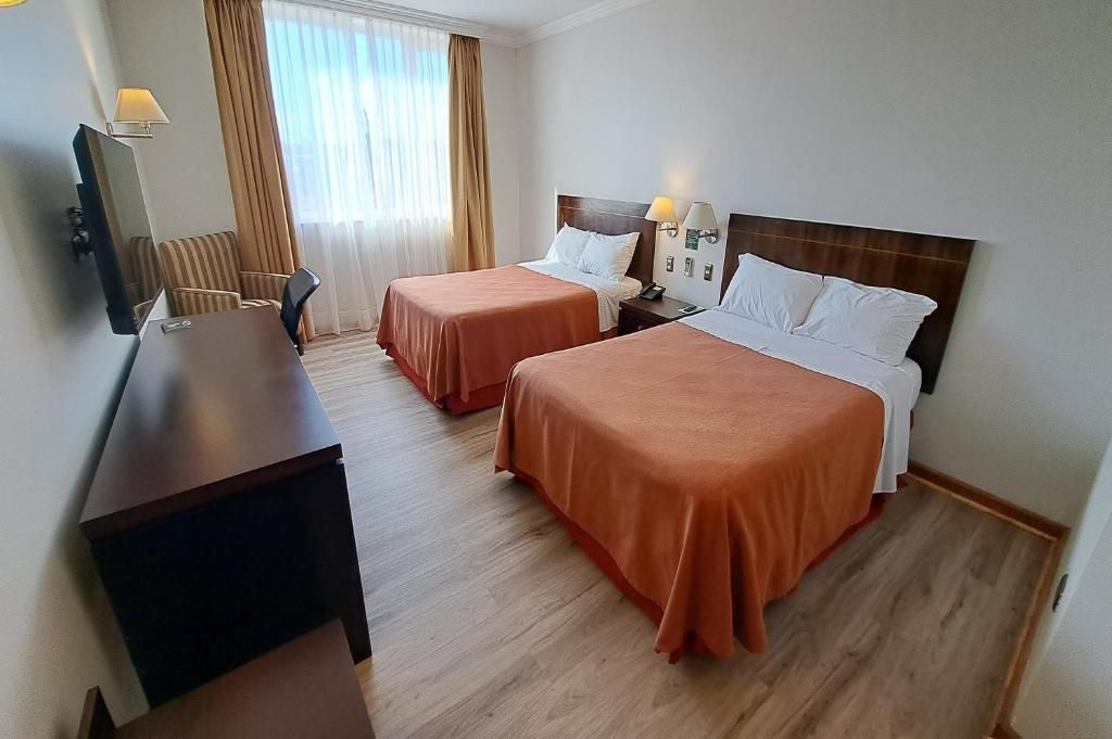 A hotel room with two beds, a desk, a chair, a TV, and a window with curtains.