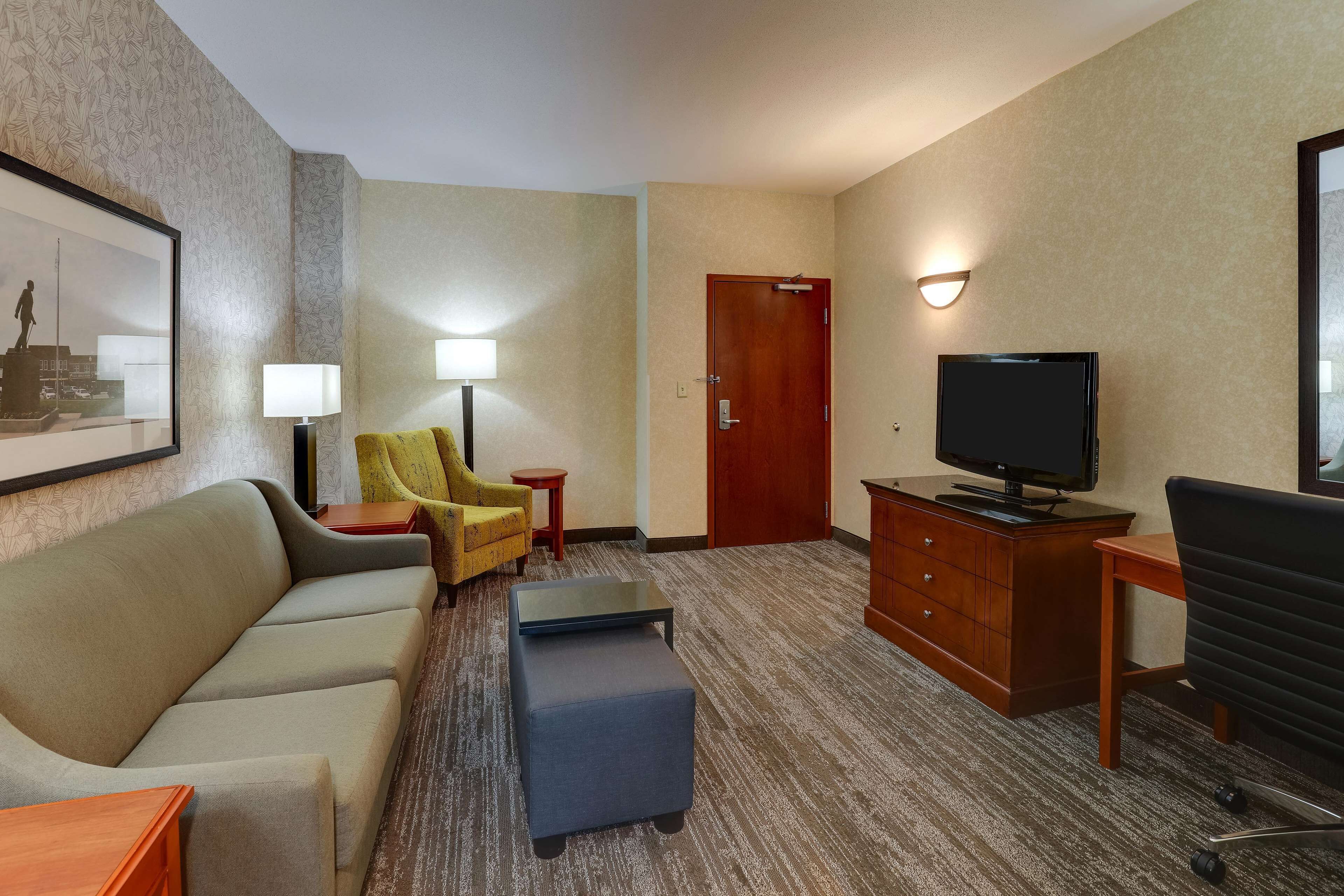 Ein Hotelzimmer mit einer Couch, einem Sessel, einem Hocker, einem Fernseher und einem Schreibtisch.