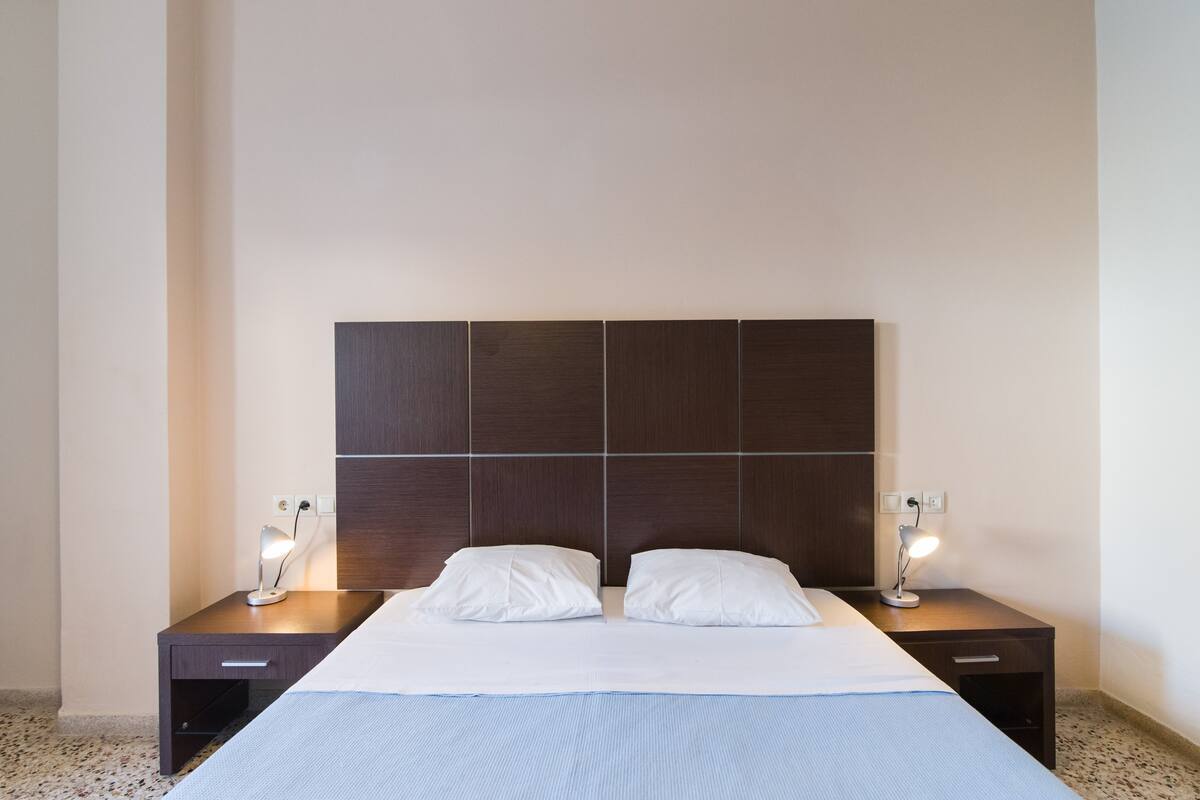 Una cama bien hecha con sábanas blancas, dos mesitas de noche con lámparas y un cabecero de madera oscura.