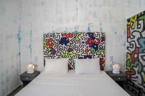 A bed with white bedding, two bedside tables with white lamps, and a colorful abstract wall pattern. - Beach Boutique Hotel (Santorini)
