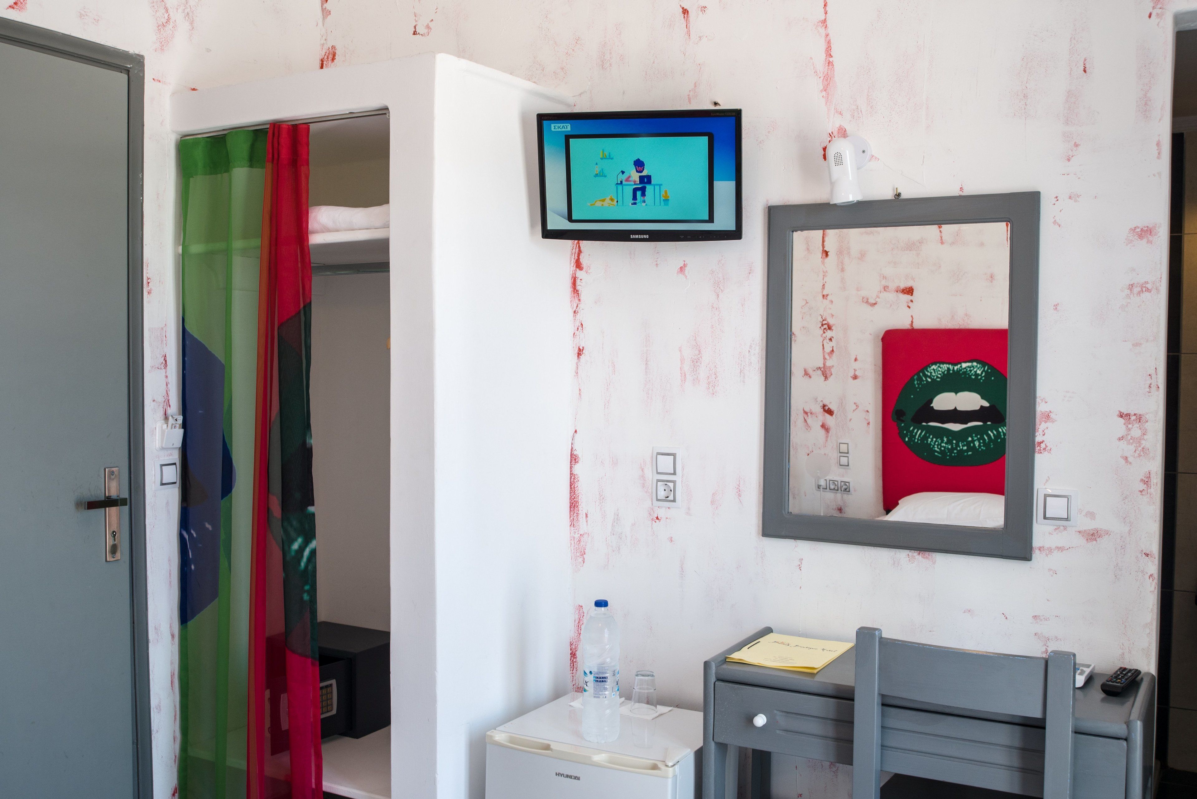 A hotel room with a television mounted on the wall, a mirror with a red artwork, a small desk with a chair, and a water dispenser.