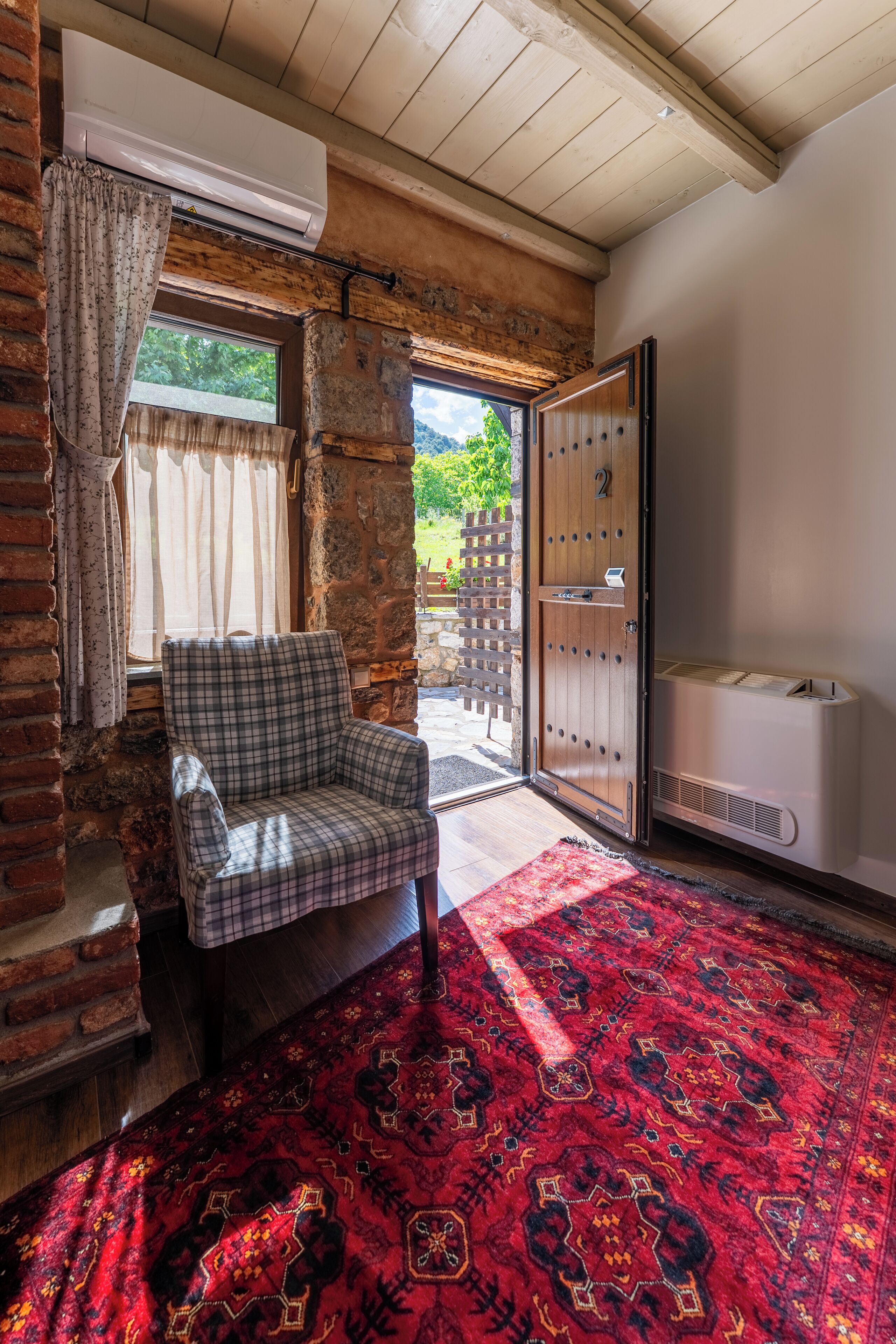 Una habitación con una silla de cuadros, una alfombra roja con estampado, una puerta de madera y una ventana con cortinas.