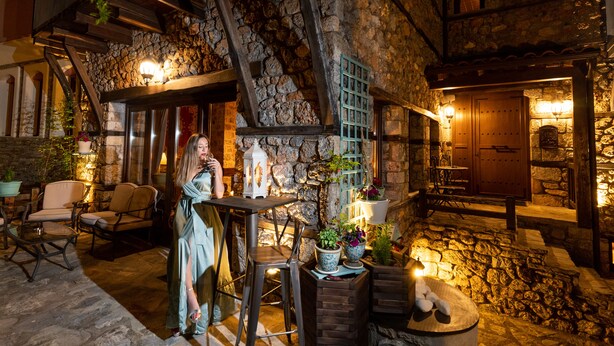 A stone building with a wooden staircase, outdoor seating, and a woman standing by a table. - To Palio Litohoro (Dio-Olympos)