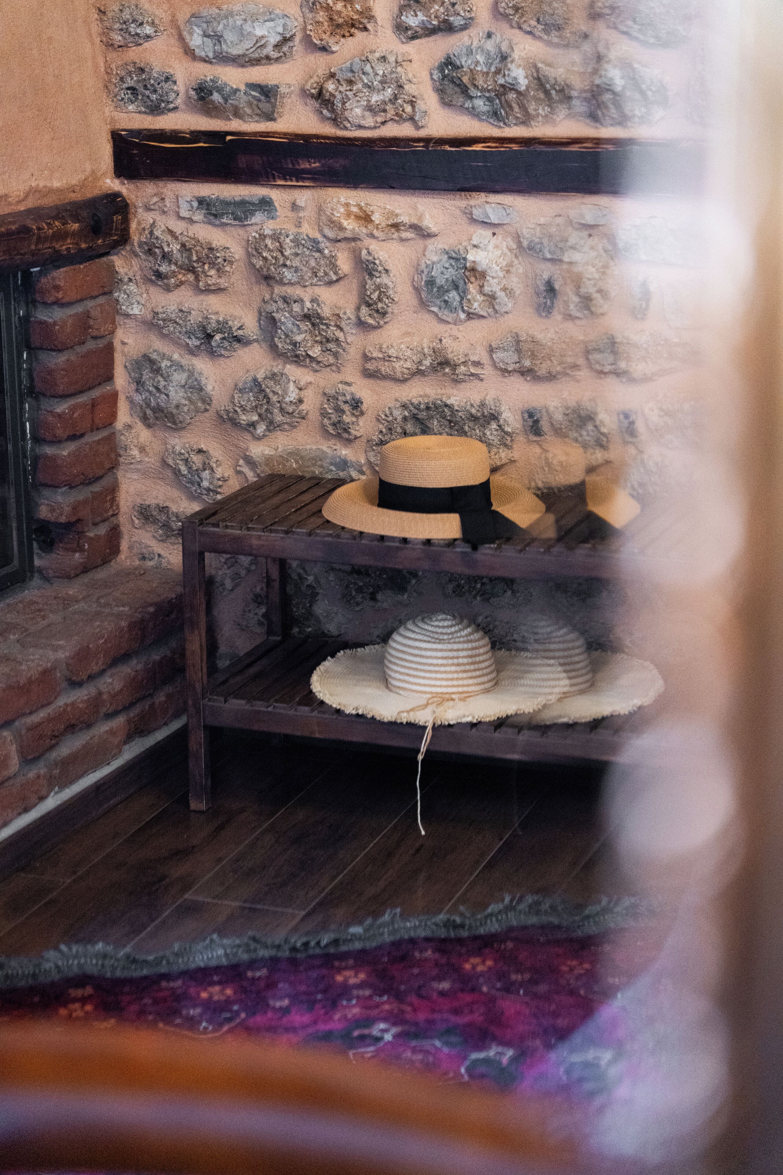 A rustic interior with a stone wall, a wooden table with hats, and a brick fireplace.