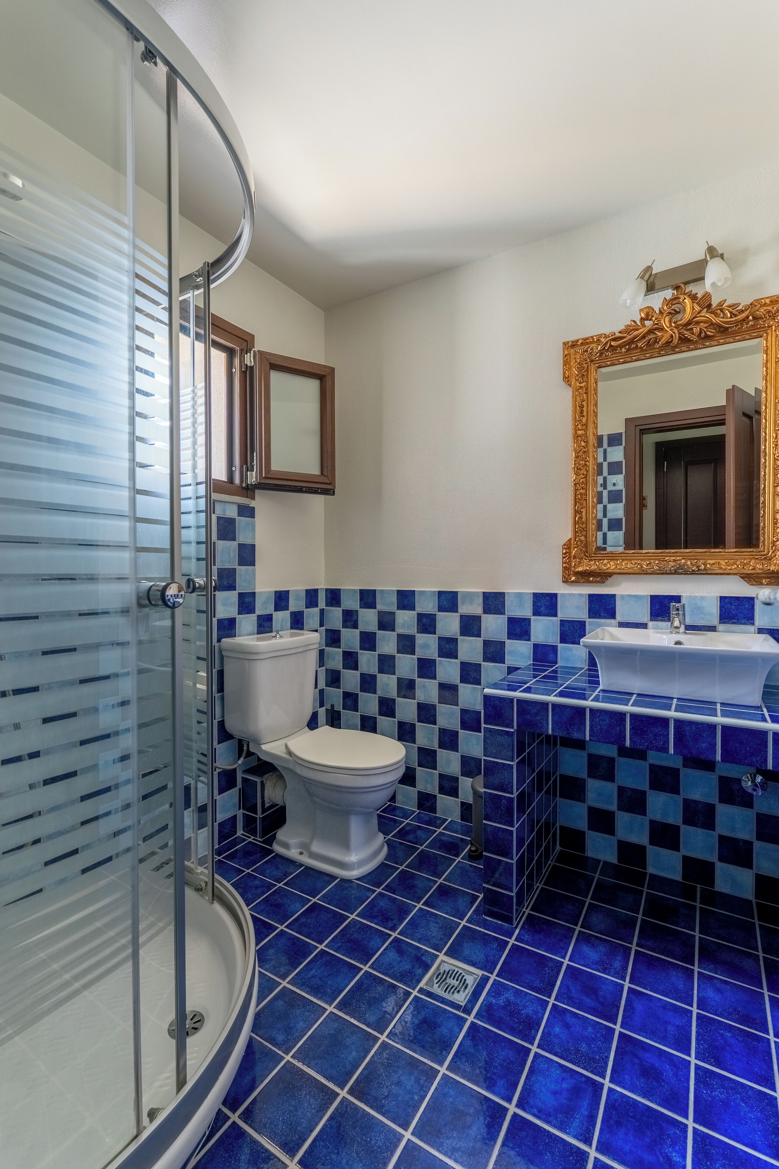A bathroom with a glass shower enclosure, a white toilet, a sink with a gold-framed mirror, and blue checkered tiles on the walls and floor.