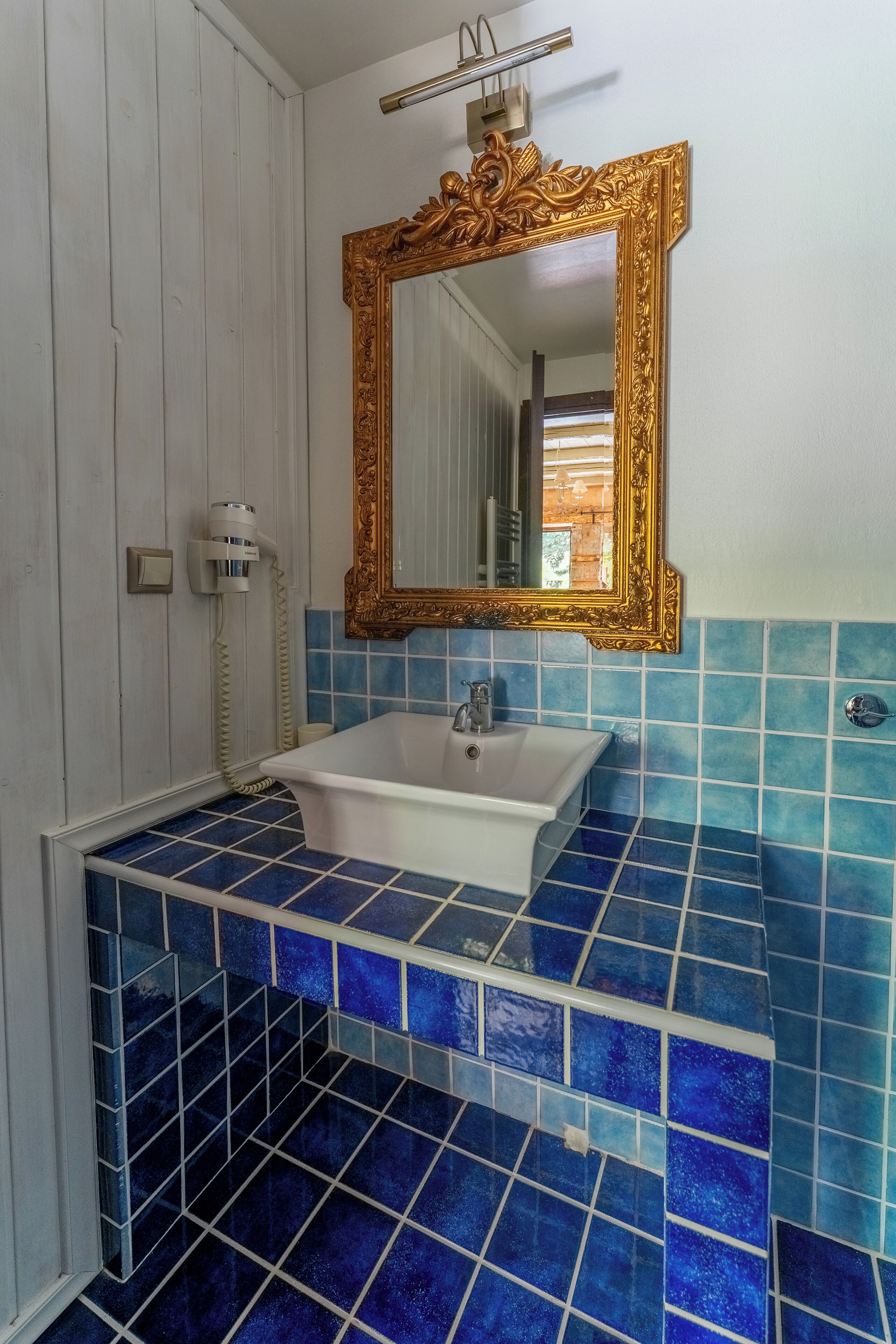 A bathroom with a white sink, blue mosaic tiles, a gold-framed mirror, and a white door.