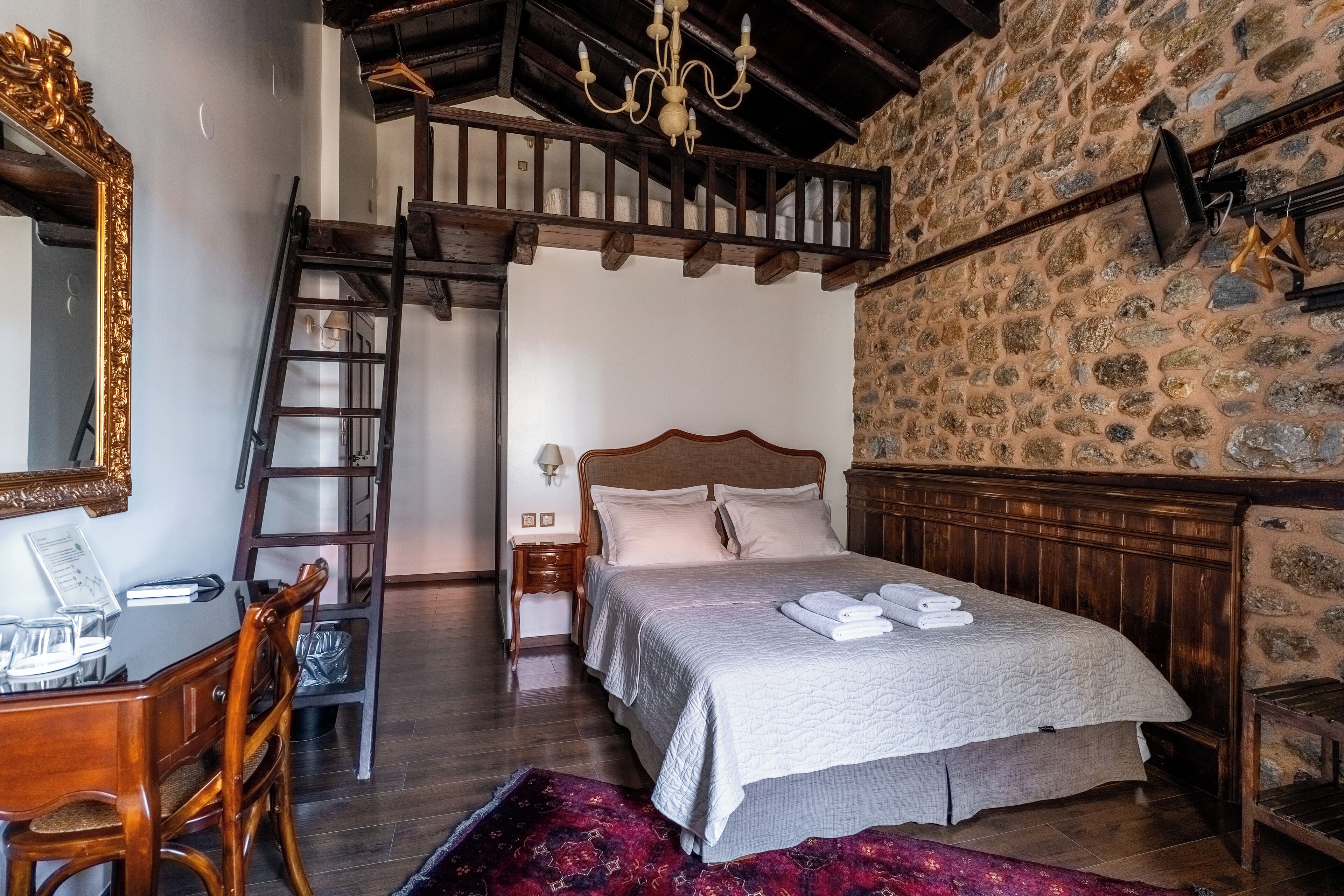Un dormitorio con una cama de madera, un escritorio con silla, una escalera, una araña y una pared de piedra.