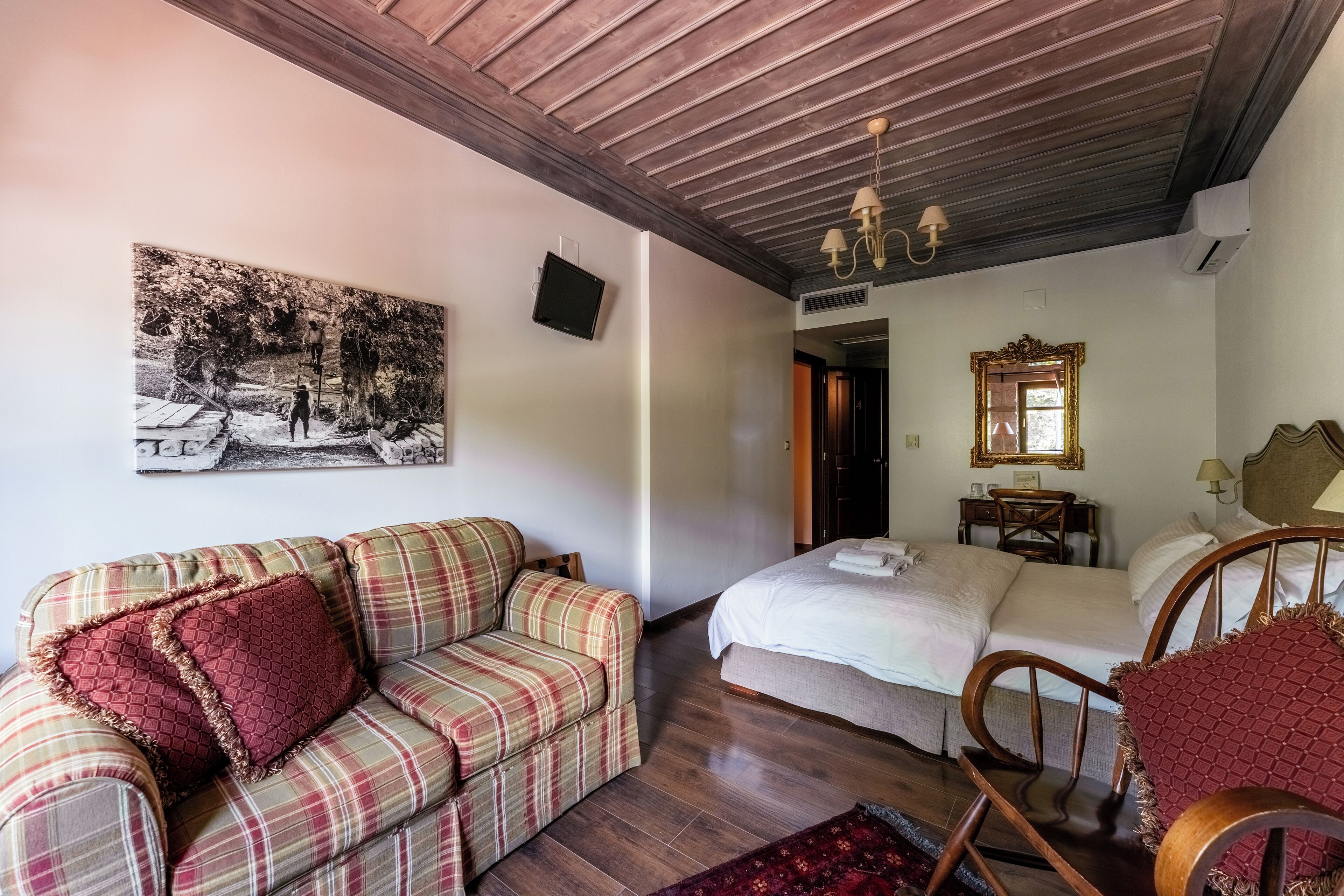 Una stanza con un divano a quadri, un letto con lenzuola bianche, un soffitto in legno e un quadro appeso al muro.