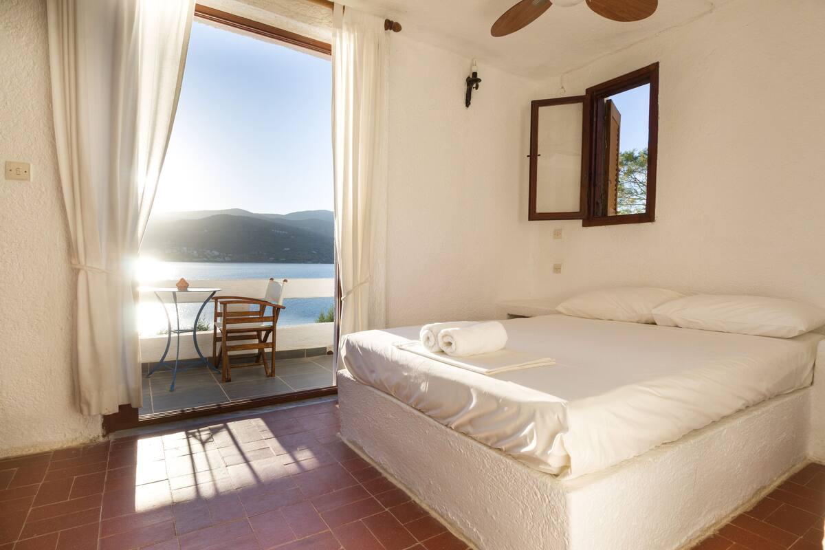 A bedroom with a bed, a chair, a table, and a view of the sea.