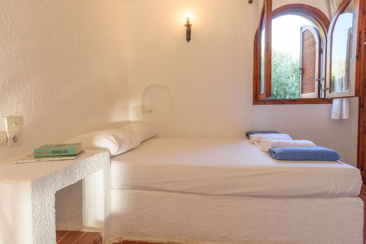 A room with a bed, a bedside table, a wall-mounted light, and a window with an arched design.