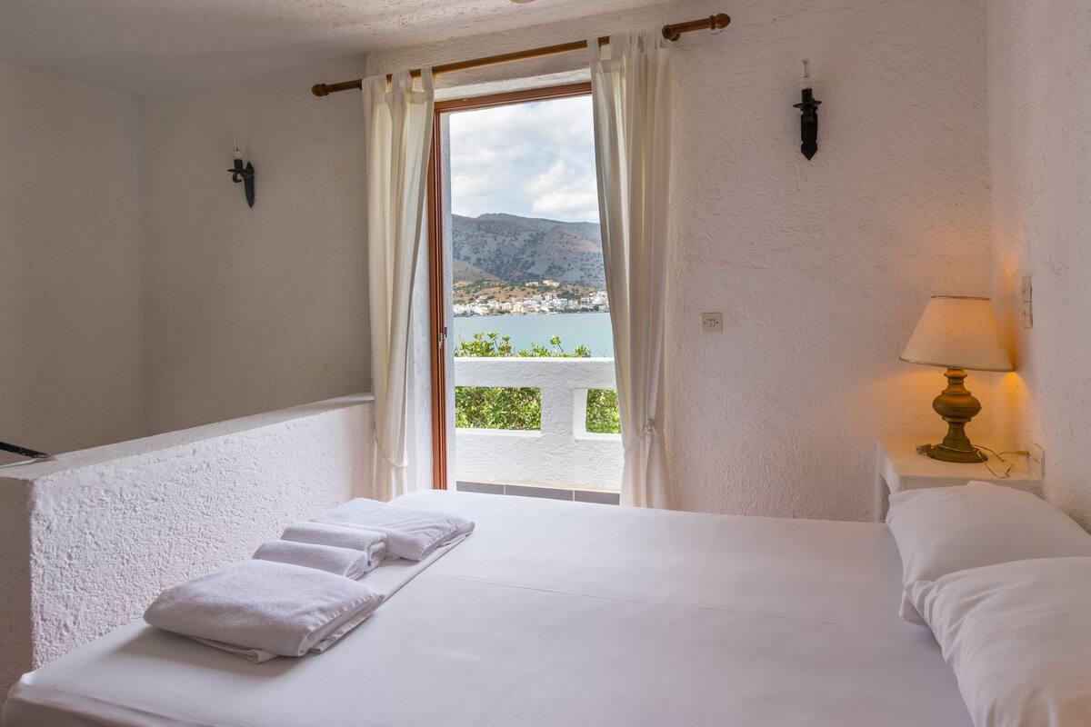 A bedroom with a bed, a nightstand, a lamp, a window with curtains, and a view of a town and mountains.
