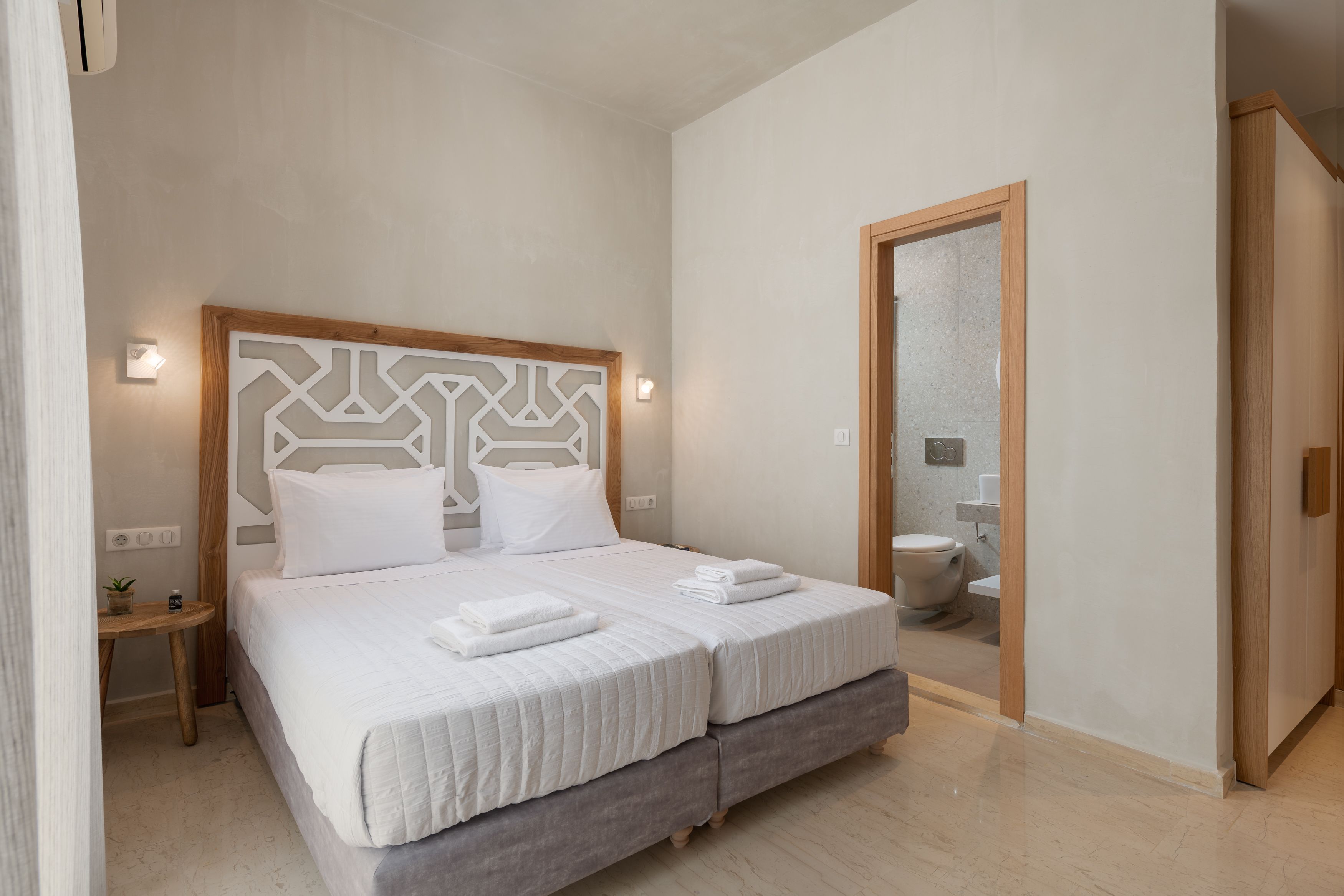 Cama doble con cabecera de madera, mesita de noche con una planta, y un baño visible a través de una puerta abierta.