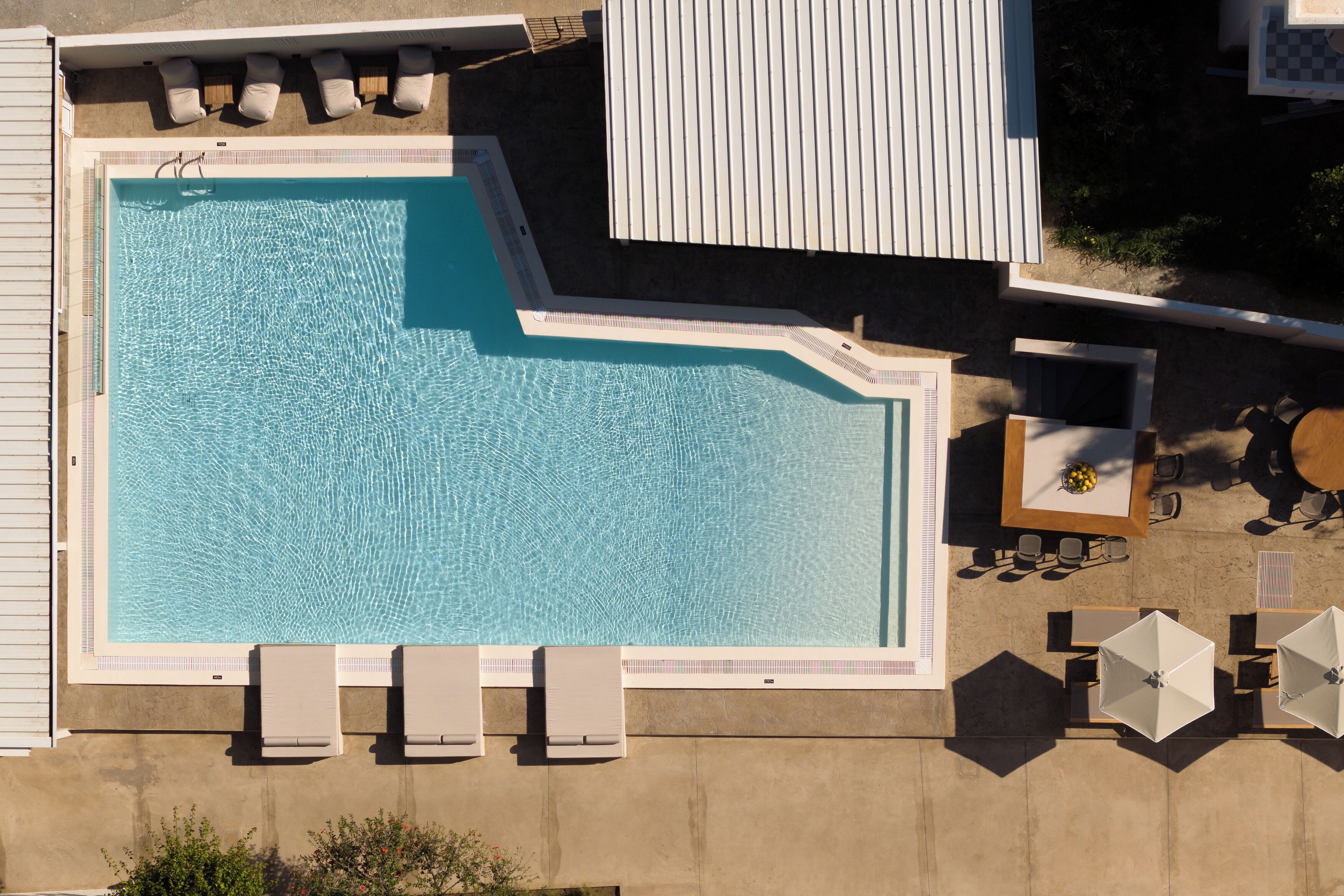 Vista aérea de una piscina con tumbonas y sombrillas, rodeada por un edificio con techo de tejas y un pequeño jardín.