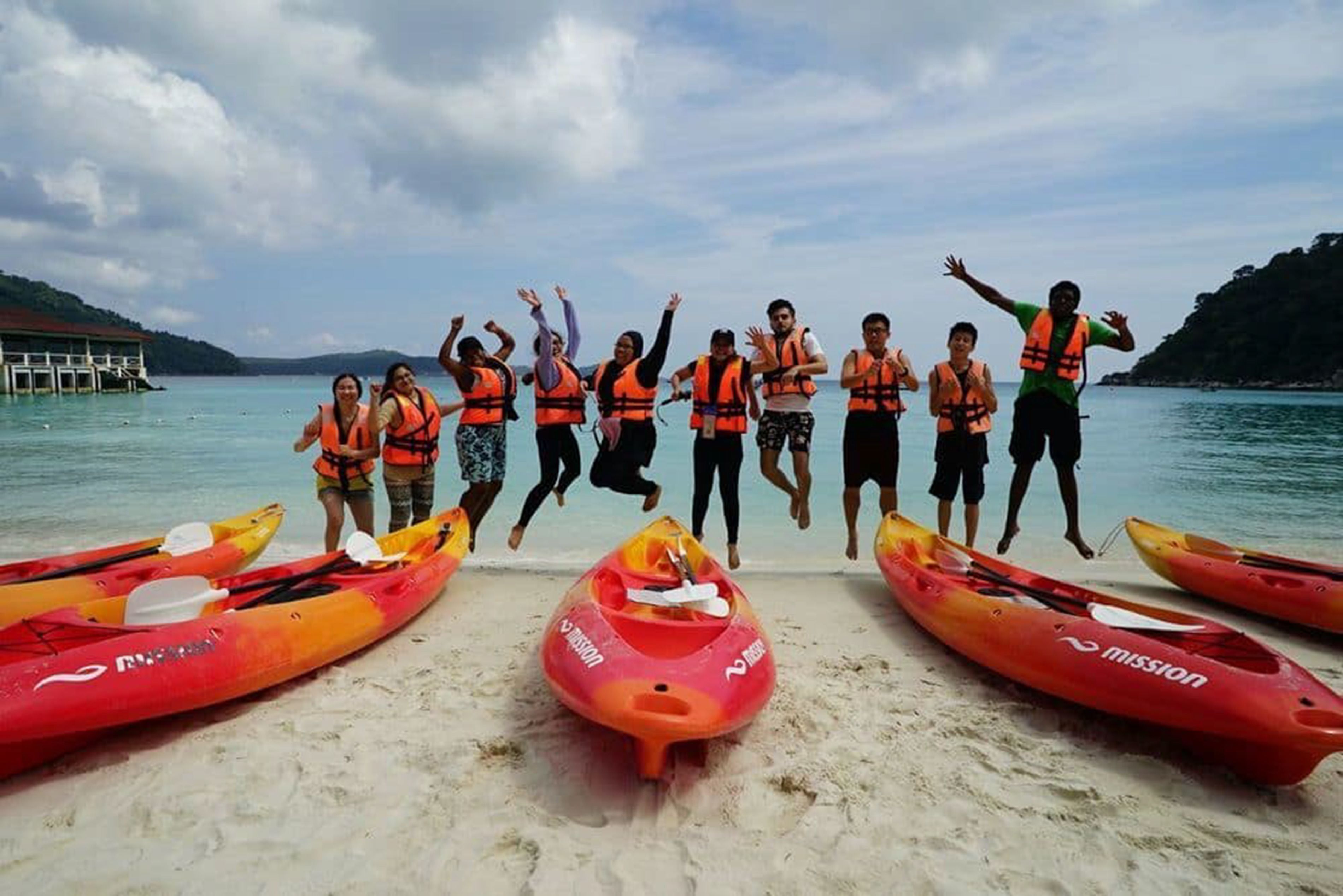 A group of people in life jackets jumping in front of red kayaks on a beach with calm water and a dock in the background.