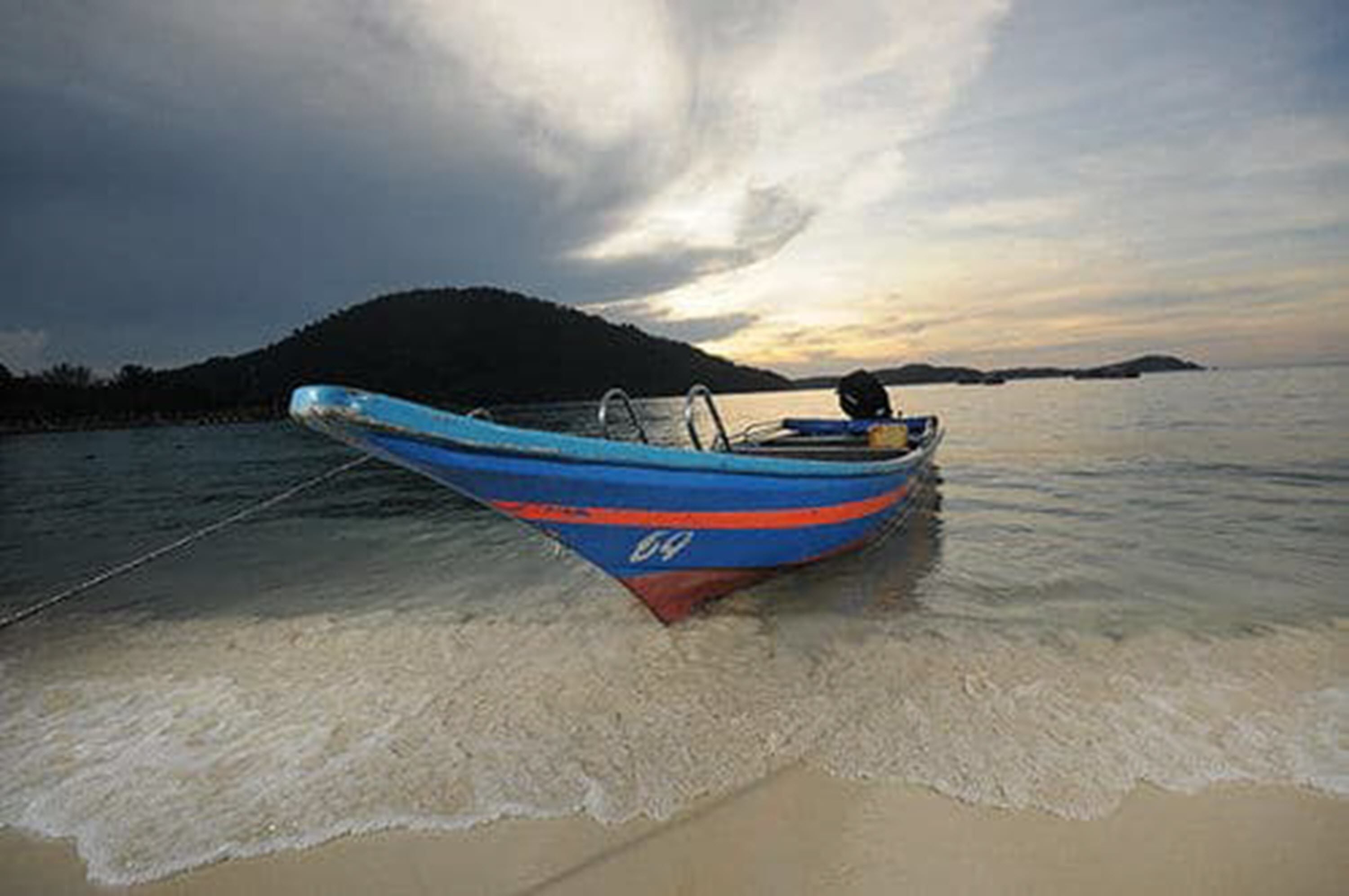 A boat with a blue and red stripe, anchored on a sandy beach with calm water and a hilly landscape in the background.