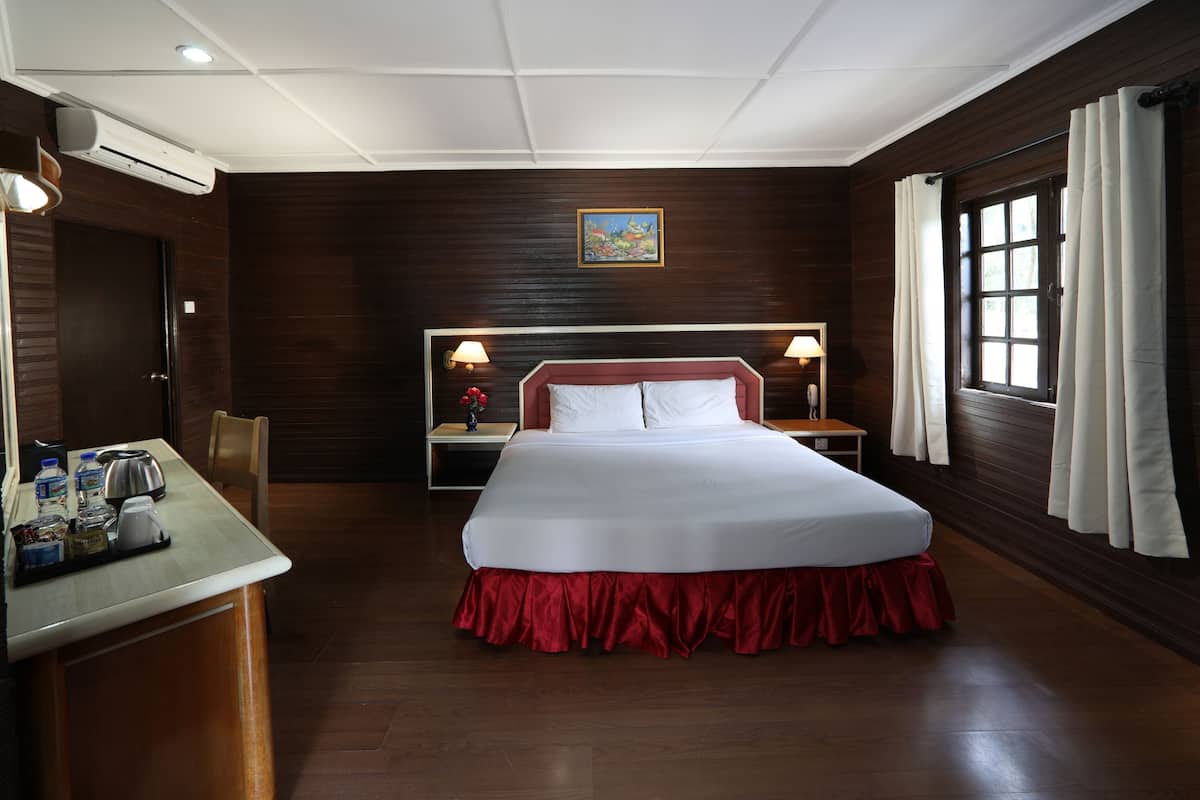 A hotel room with a large bed, a desk with a kettle, a nightstand with a lamp, and a window with curtains.