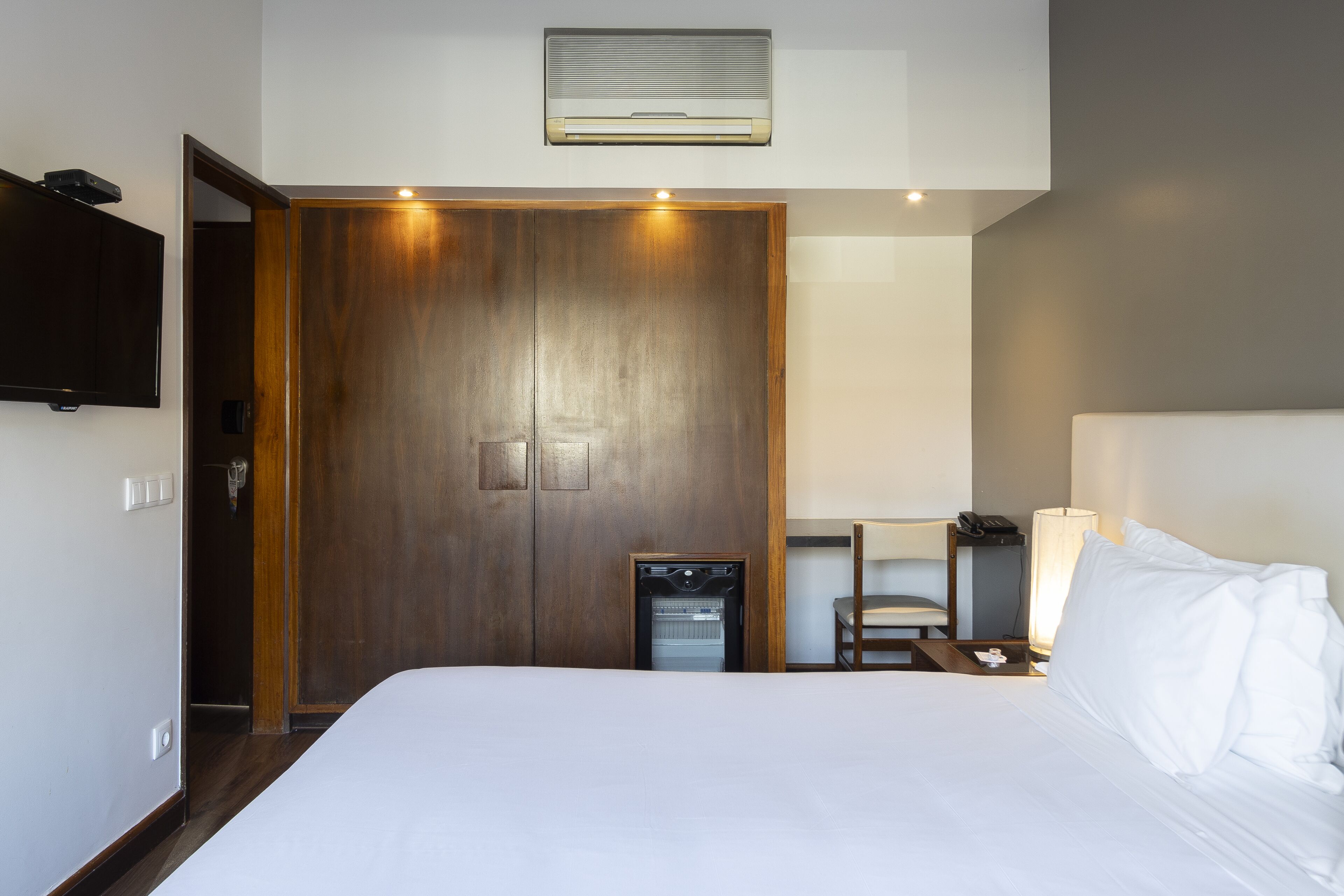 Una habitación de hotel con una cama grande, un armario de madera, un escritorio con lámpara y un refrigerador pequeño.