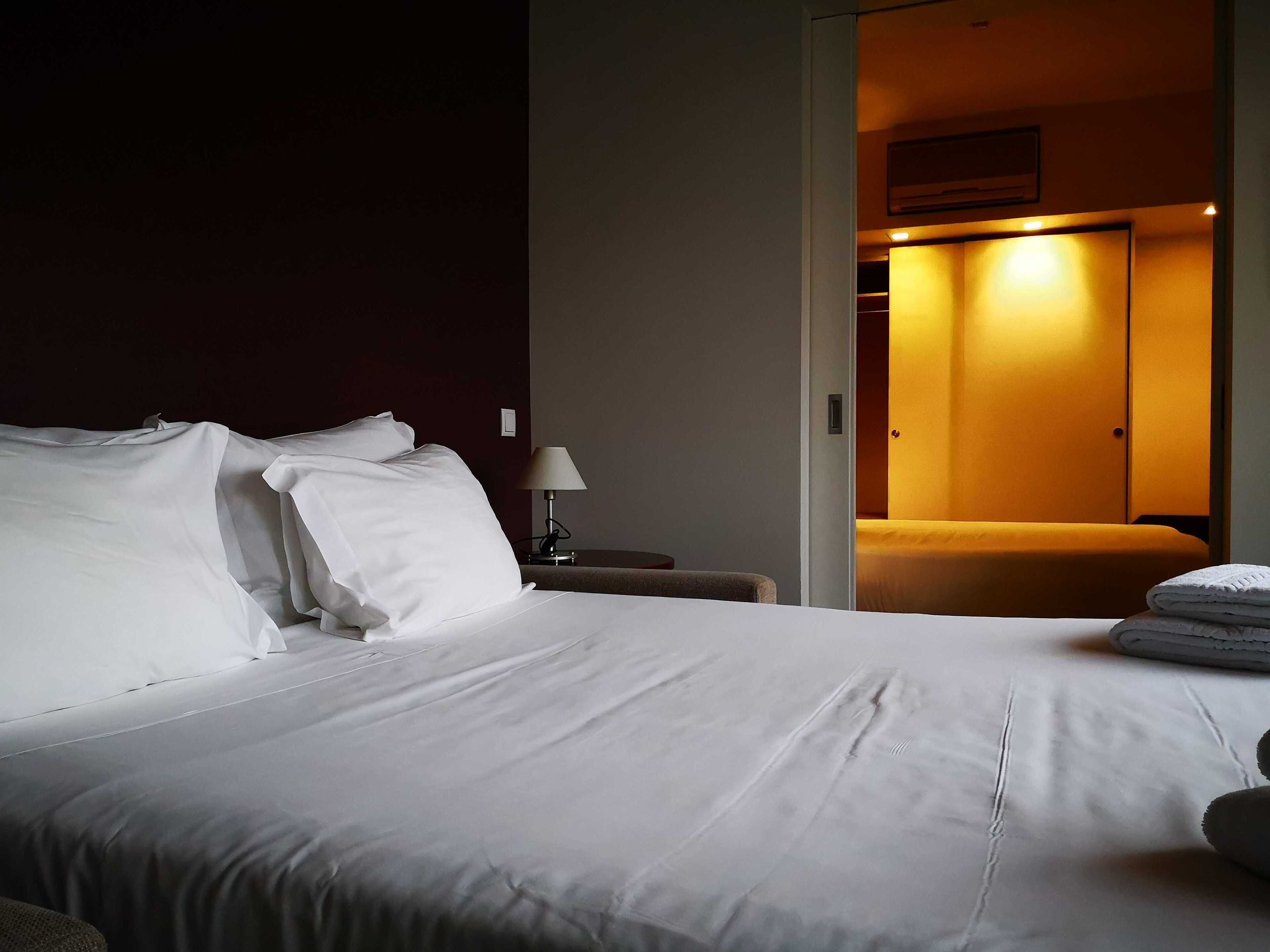 Una habitación de hotel con una cama grande, una mesita de noche con lámpara y una puerta que da a un baño.