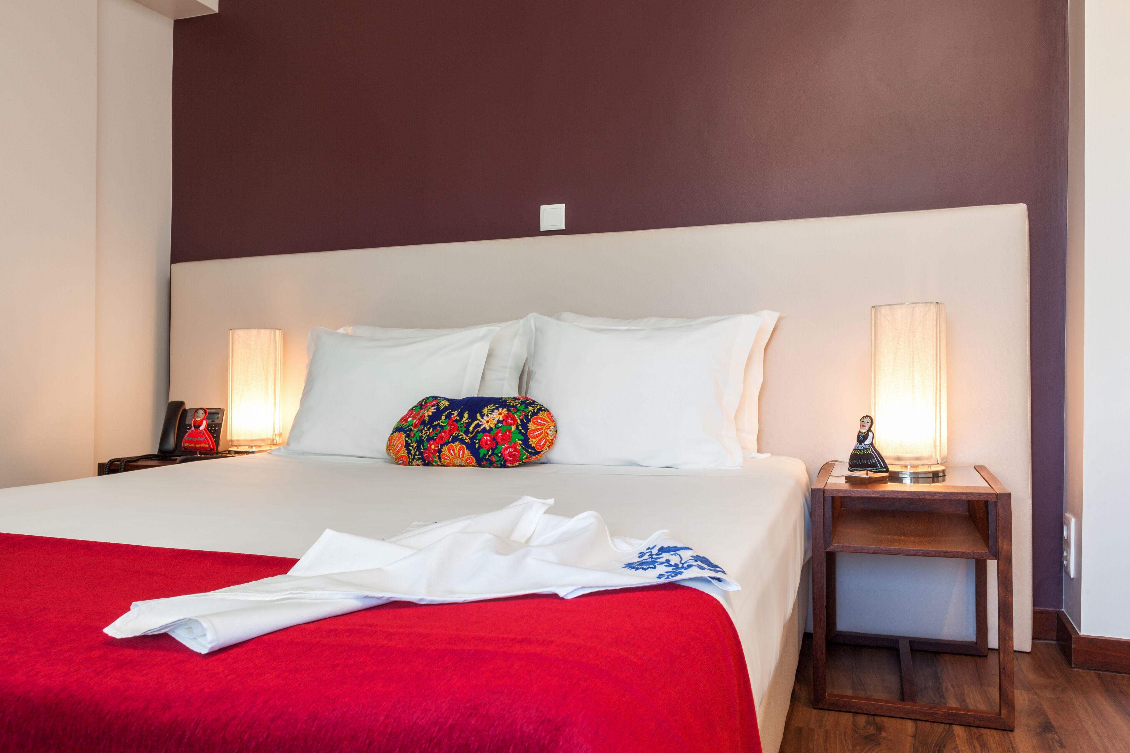 Een net opgemaakt bed met een rode deken, witte kussens en een kleurrijk sierkussen. Aan beide zijden van het bed staan nachtlampjes.